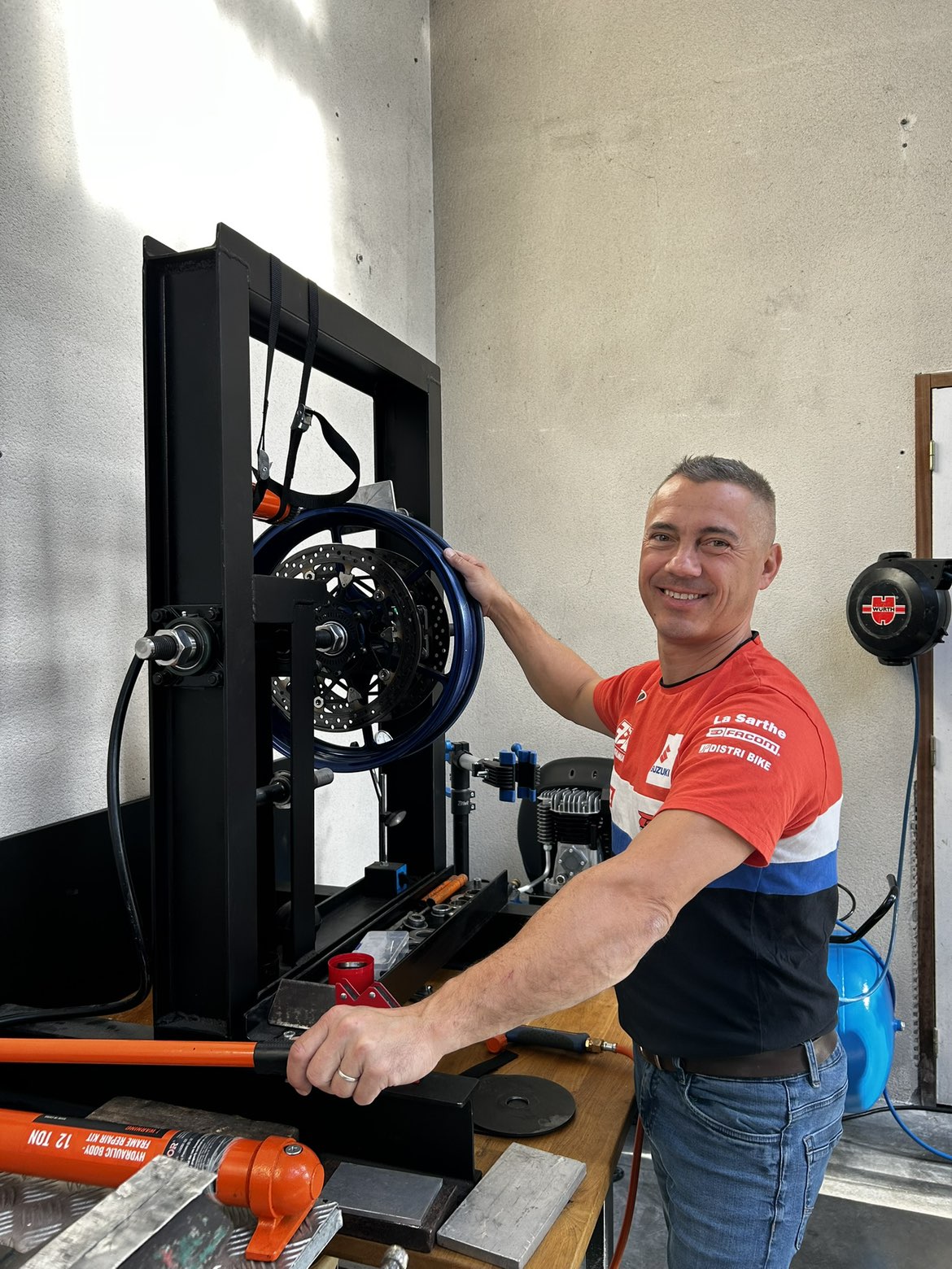 Mécanicien de ROUBY SEB MOTO souriant à côté d'une jante bleu foncé à double disque montée dans un banc de redressage hydraulique, kit de réparation 12 tonnes orange sur l'établi.