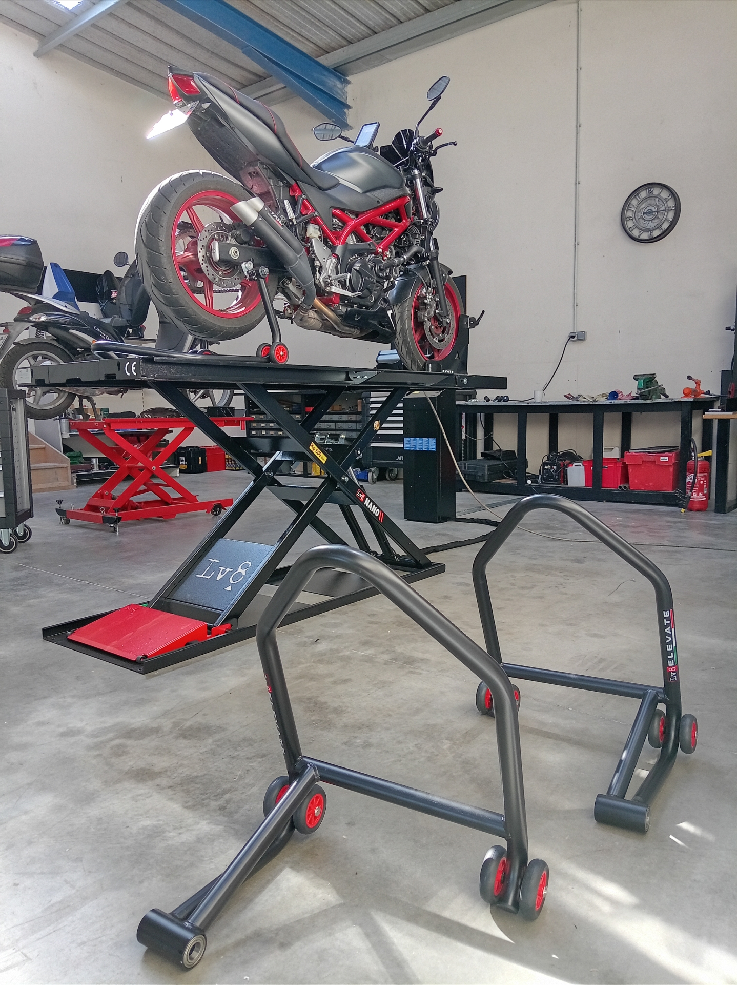 Moto naked rouge et noire sur pont élévateur LV8 à l'atelier, béquilles d'atelier au premier plan, établi et matériel de mécanique en arrière-plan.