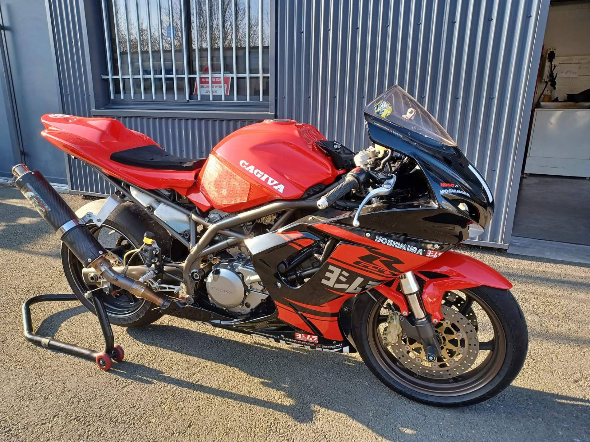 Prototype piste sur base de Cagiva Raptor rouge posé sur béquille d'atelier devant l'atelier : carénage Suzuki GSX-R, kit déco Yoshimura, réservoir rouge marqué CAGIVA, silencieux Yoshimura carbone.